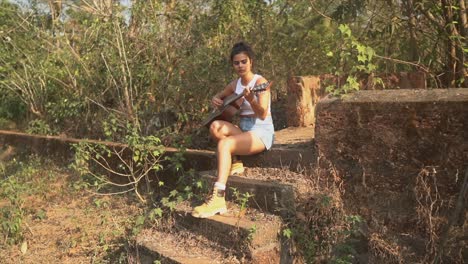 Brunette-girl-playing-a-guitar-sitting-on-some-concrete-steps-on-a-stage-with-dry-vegetation