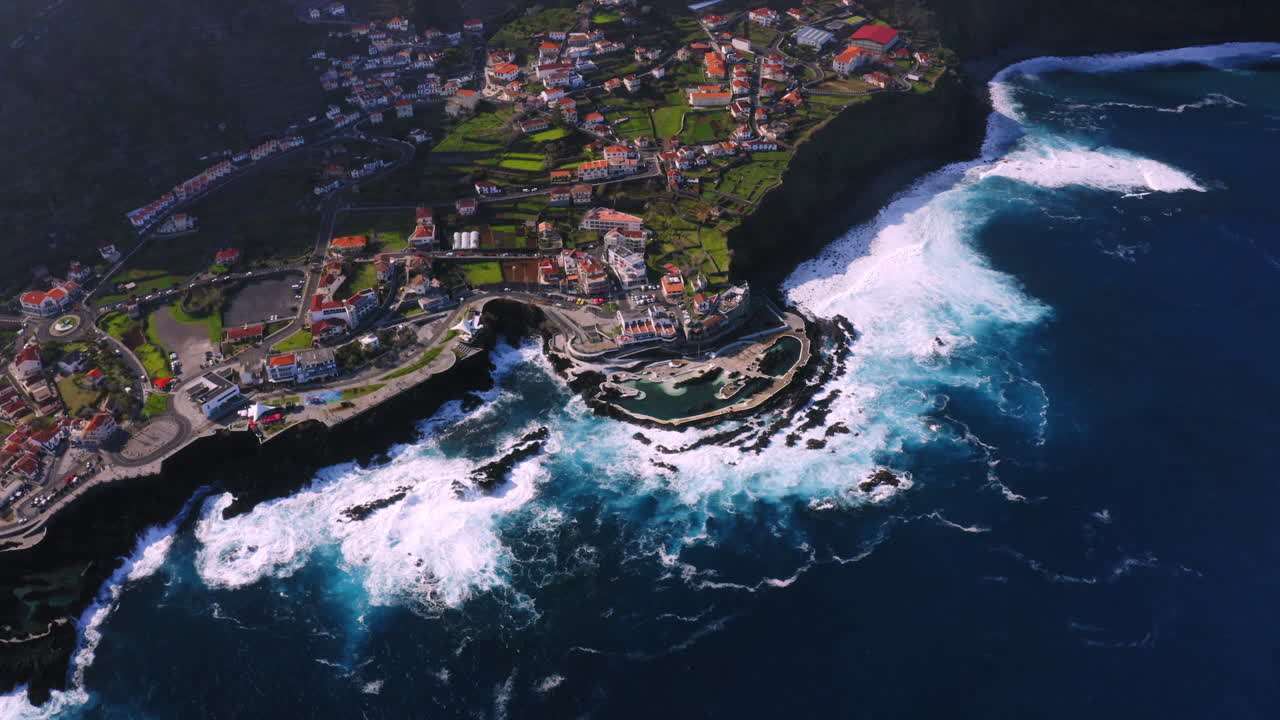 Vista aerea in discesa di Porto Moniz con piscina naturale e onde che si infrangono contro la costa durante una giornata di sole
