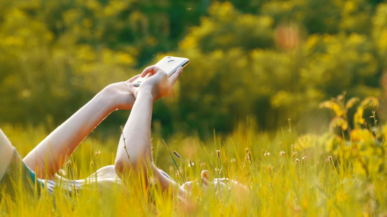 Happy Young Woman Lying on Green Lawn Uses Smartphone on Scenic Field at Sunset Background