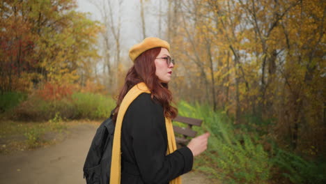 side view of tourist in black coat, yellow scarf, and beret stepping into wooden gazebo in serene autumn park, adjusting hair while holding bag strap