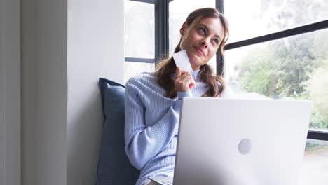 Woman shopping online with credit card, smiling and sitting by window at home