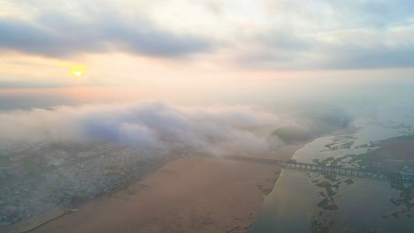 Aerial drone shot of Vijayawada city waking up to the first rays of the sun, with clouds forming a soft blanket below.