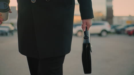 close-up of man in black outfit walking outdoors with briefcase in left hand and coffee in right hand, blurred background features parked cars