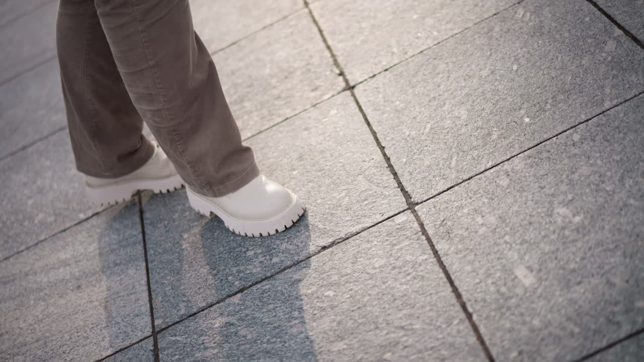 Closeup of girl legs in beige corduroy pants and chunky white platform shoes tapping and gliding across snow dusted plaza tiled floor, with long evening shadows and soft pale winter sunlight