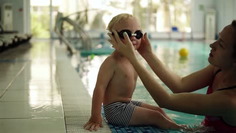 ein kleiner schwimmer bereitet sich zum schwimmen vor. der junge sitzt am rand des pools, seine mutter setzt seine schwimmbrille an. er schwenkt mit den füßen auf dem wasser. drinnen