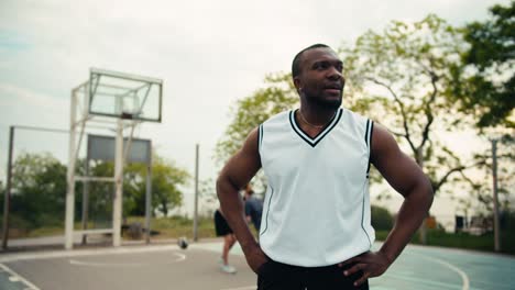 Dynamic-portrait-of-a-Black-skinned-man-who-is-standing-in-a-white-t-shirt-on-a-basketball-card-in-front-of-his-friends-who-are-playing-basketball