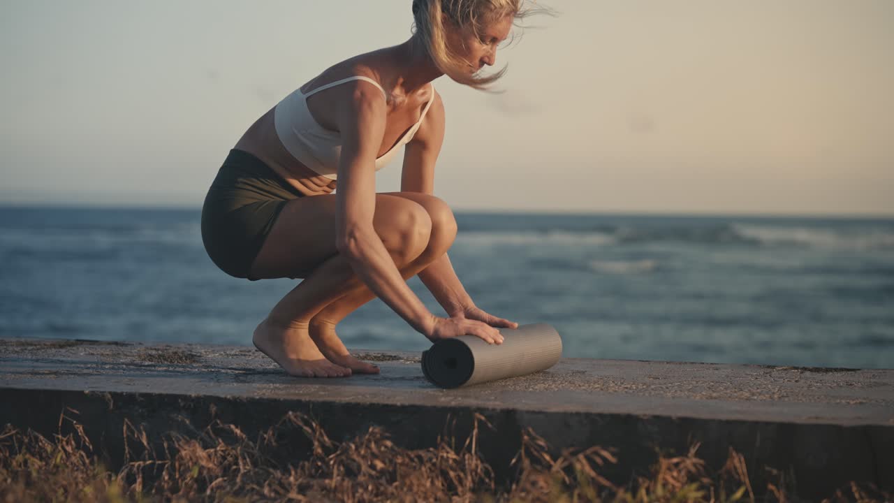 mujer en forma en ropa deportiva desplegando colchoneta de yoga en la orilla del rompeolas de hormigón, atardecer