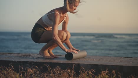 fit woman in sportswear rolling out yoga mat on concrete breakwater shore, dusk