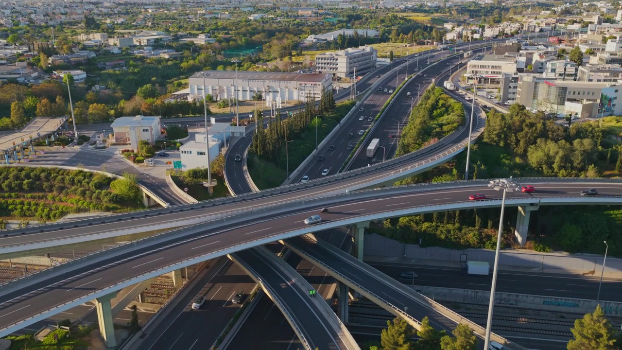 Korkealta kuvattu ilmakuva autoista ja kuorma-autoista ajamassa monimutkaisessa monikerroksisessa moottoritieliittymässä, jota ympäröivät vihreät puut ja kaupunkikehitys aurinkoisena päivänä.