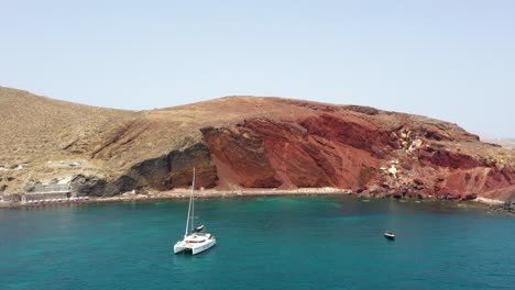 antena volando lateralmente junto a la playa roja con agua turquesa, botes, montañas y arena de color rojo en santorini, grecia