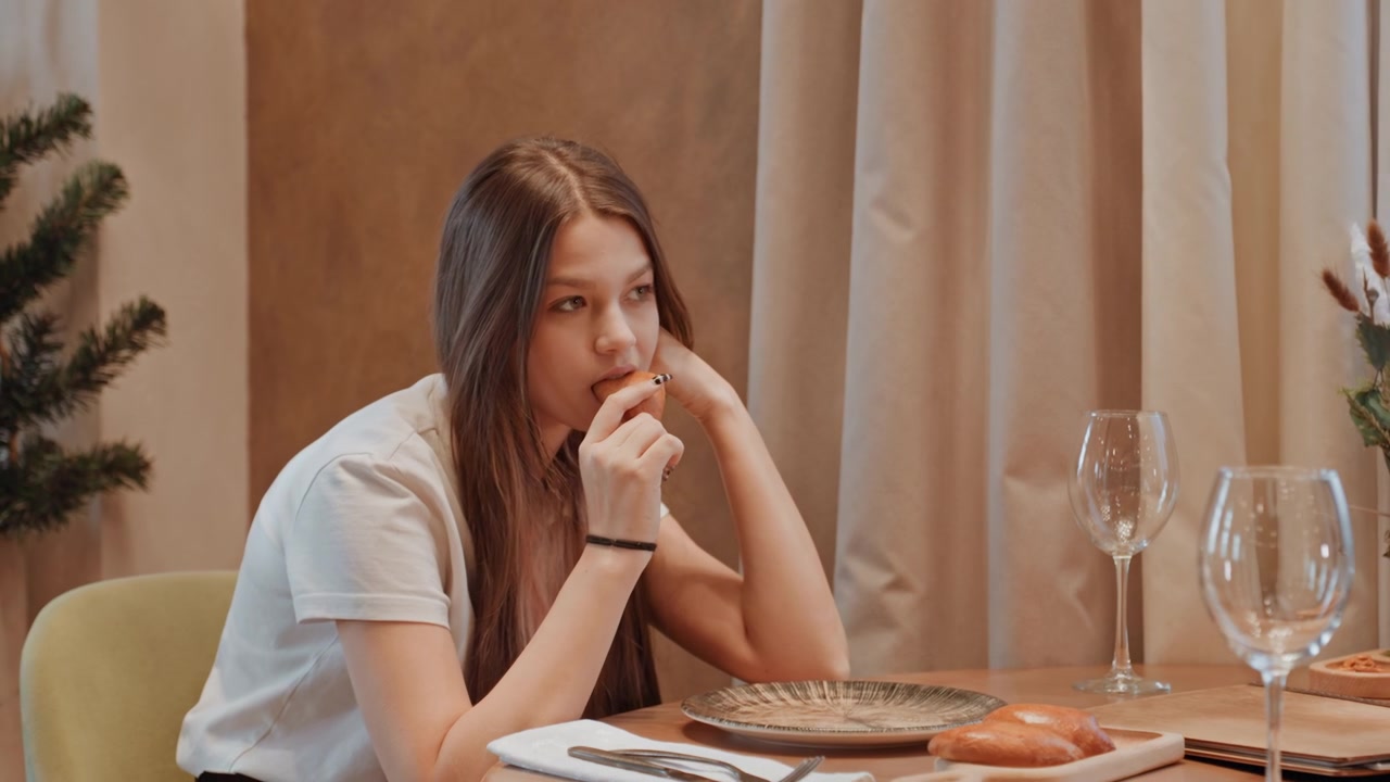 Giovane donna che aspetta da sola al tavolo, blogger che controlla il telefono con il mento irrequieto sulla mano, pasticcino intatto sul piatto, toni interni neutri e bicchieri vuoti, sottile tensione e bokeh cinematografico.