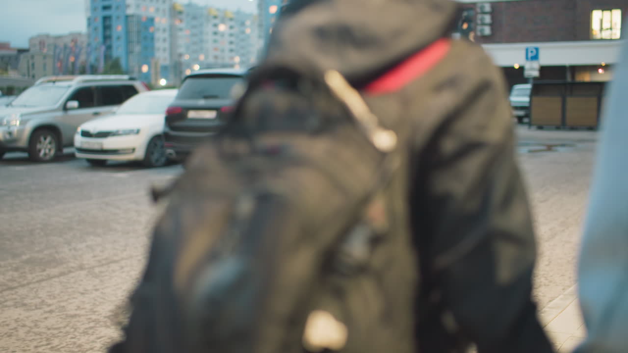 Close-up of backpack worn by student in black jacket walking next to person in denim jacket, showing daily urban routine, casual fashion, and preparation for school or travel on city street in soft evening light