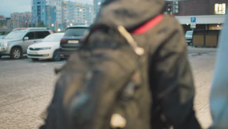 close-up of backpack worn by student in black jacket walking next to person in denim jacket, showing daily urban routine, casual fashion, and preparation for school or travel on city street in soft evening light