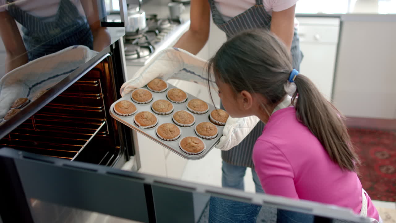 madre y hija bi-raciales felices horneando en la cocina sacando pasteles del horno, cámara lenta