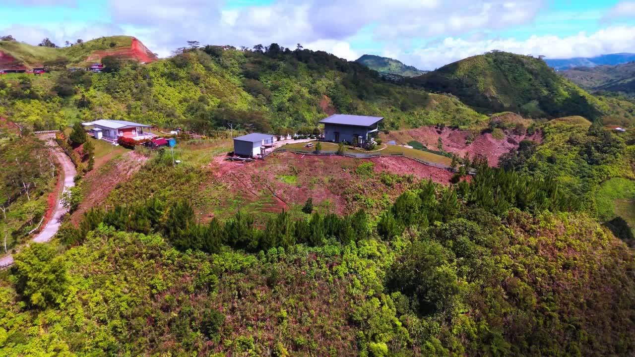 drone shot captures several modern homes perched on a grassy hilltop amid rolling, tree‑covered mountains. A winding road leads up to the houses, the scene highlights verdant landscape.The bright sky