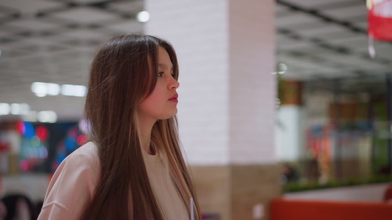 Side view of young lady with long hair sipping juice from cup while standing in modern mall setting, casual outfit with relaxed expression, blurred background