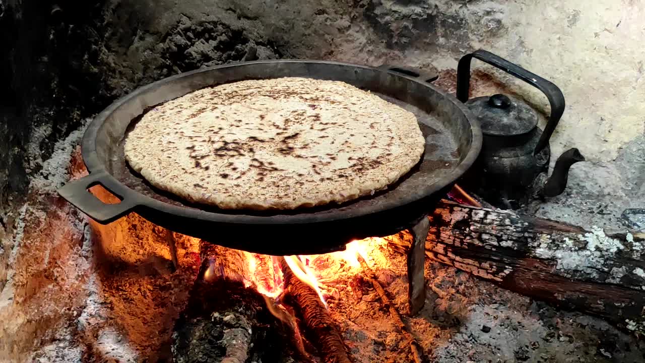 primer plano de cocinar pan en una sartén de hierro fundido descansando sobre salvamanteles, sobre un fuego de madera de roble en una gran chimenea de piedra en una cocina tradicional-1