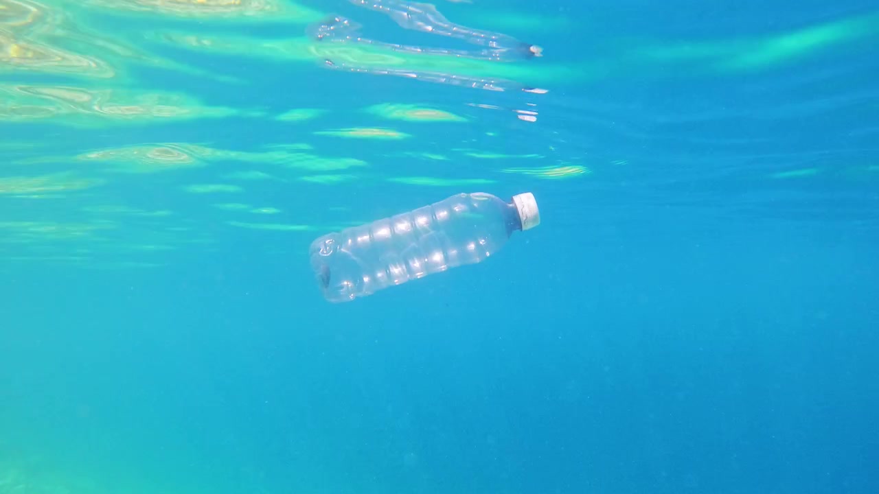 Una bottiglia di plastica è sospesa sott'acqua in acqua oceanica limpida. La luce del sole filtra, illuminando la bottiglia e l'acqua circostante. Sotto è visibile una barriera corallina.