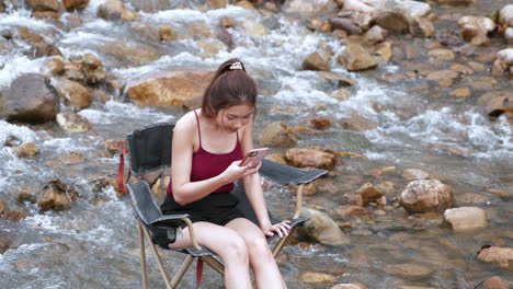 Mujer-Asiática-De-Rojo-Sentada-En-Una-Silla-Con-Grandes-Rocas-Y-Agua-Salpicando-Lentamente-Para-Relajarse-Y-Ser-Feliz-En-Las-Vacaciones-De-Verano