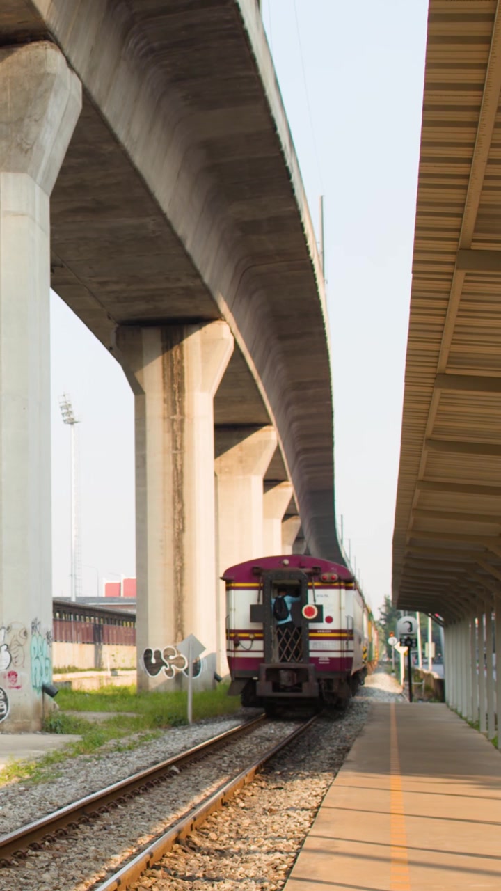Kereta penumpang berangkat dari peron stasiun di bawah pilar beton besar pada siang hari.