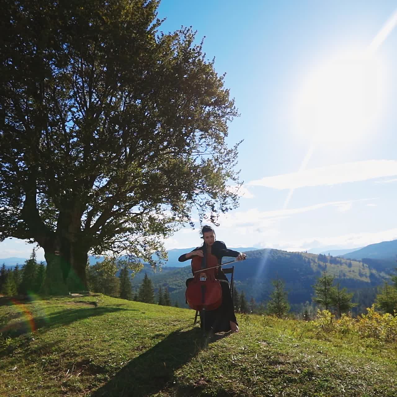 Dark-haired woman in black long dress playing cello. Lady musician making classic music sitting on the chair in the mountainous landscape