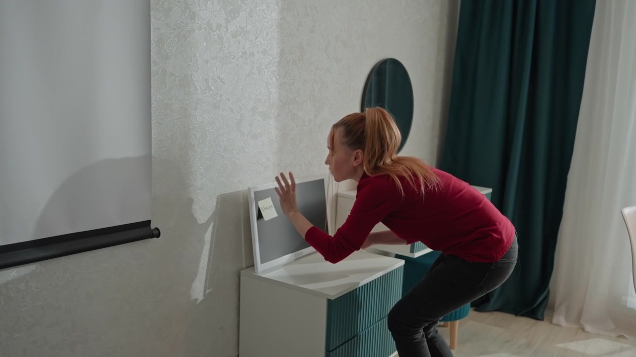 Young woman in red top and dark pants cleans electronic equipment. Woman interacts closely. Setting includes white sheer curtains green drapes round mirror showing woman