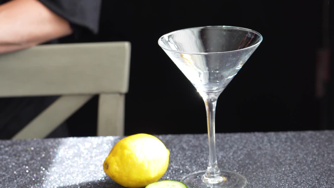 Barman si prepara a fare un cocktail in un bicchiere da Martini con fette di limone e cetriolo, usando pinze per ghiaccio e uno shaker Cobbler argentato. Primo piano delle mani di una donna con smalto rosa su un tavolo di granito.