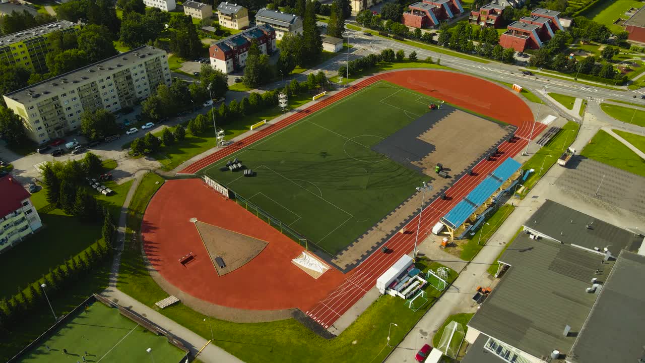 Aerial view orbiting around a colorful soccer stadium that is being built or renovated by workers with high visibility jackets and machines, summer sunny day at a small urban town called Laagri