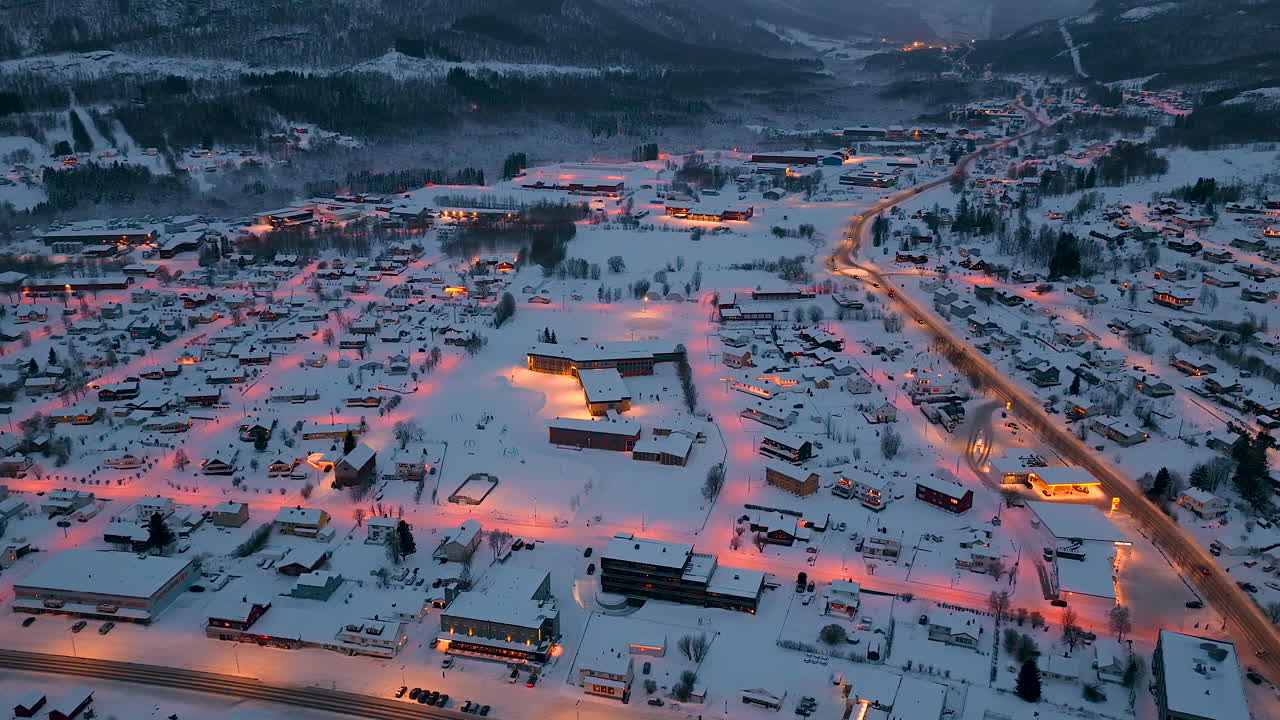 Upea ilmakuva dronella lumipeitteisestä norjalaisesta kylästä Lofoottien saaristossa sinisen hetken aikaan, lämpimien katuvalojen hehkuessa talvimaisemaa vasten.