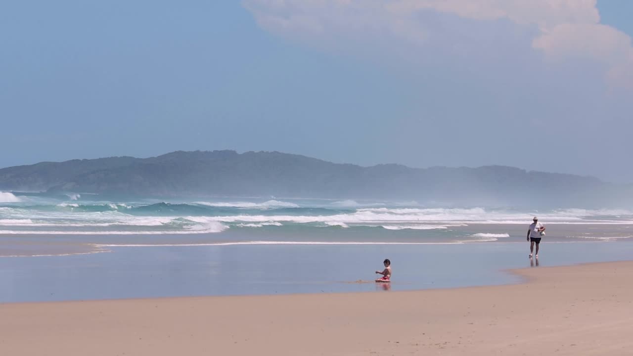 A serene beach scene with two people walking along the shore, distant hills in the background.