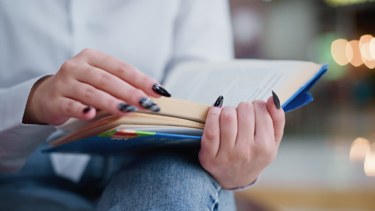 primer plano de una mujer con uñas negras hojeando las páginas de un libro abierto mientras está sentada con las piernas cruzadas, las suaves luces bokeh crean un ambiente cálido en el fondo