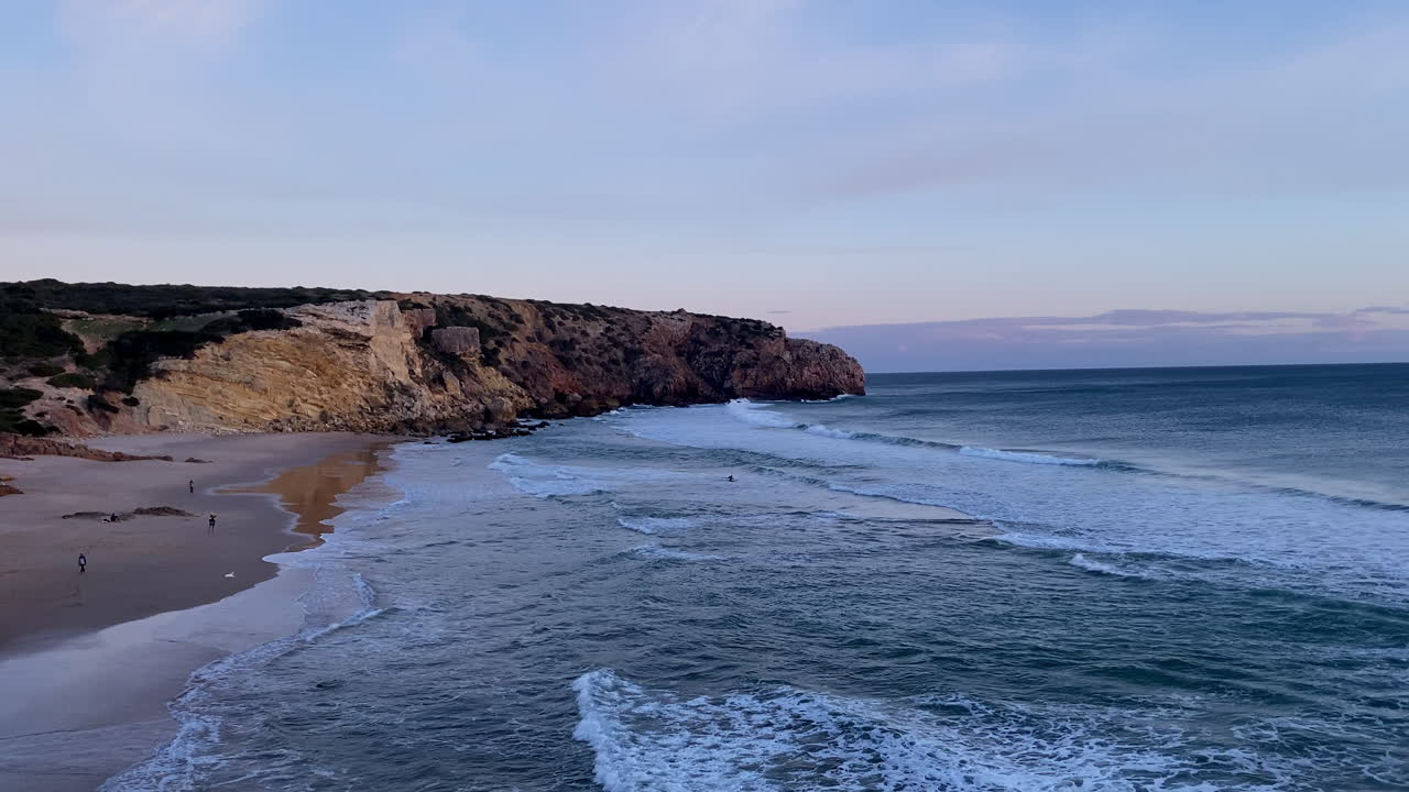 Scogliere costiere si affacciano sulle onde dell'oceano con surfisti che pagaiando al tramonto. Surfisti che pagaiando sotto imponenti scogliere al tramonto vicino alla spiaggia di Zavial, dove le onde dell'Atlantico e la luce morente definiscono l'energia grezza di
