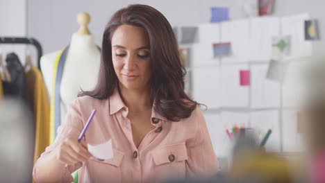 female fashion designer at desk in studio drawing designs and choosing colour and fabric swatches