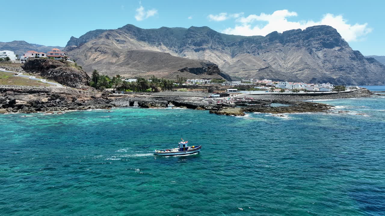 maravillosa toma aérea siguiendo un barco de pesca en las aguas del puerto de agaete y donde se pueden ver las montañas y la costa