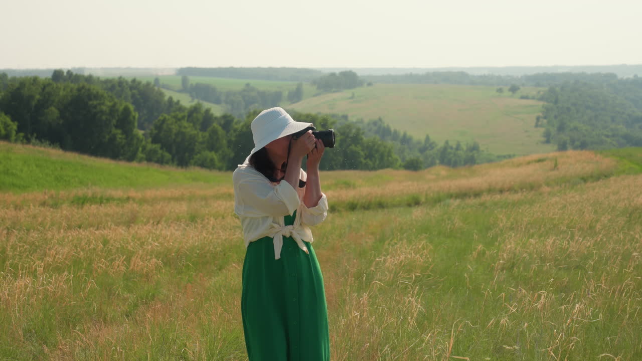 Smiling tourist wearing white hat and green dress holds camera while walking through vibrant grassy field surrounded by lush hills and distant trees under warm sunlight
