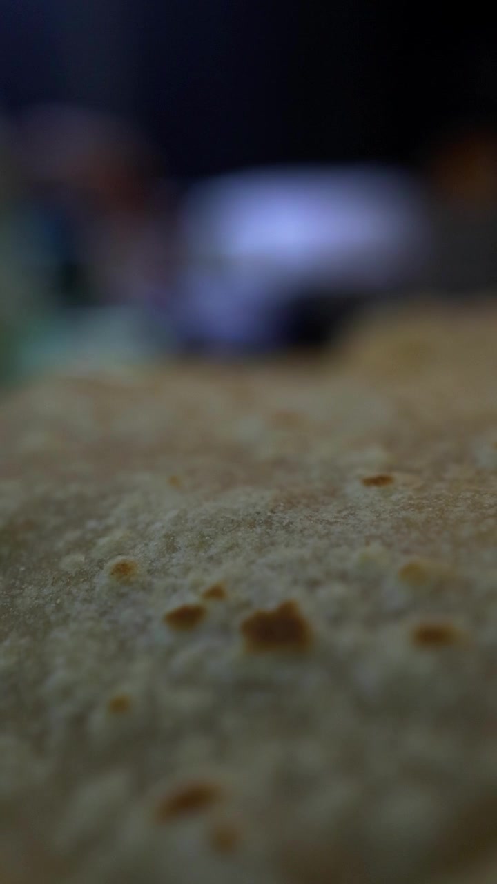 Extreme close-up of uncooked chapati dough surface showing texture and small holes, with blurred figure of cook in background preparing Indian flatbread in low indoor lighting.