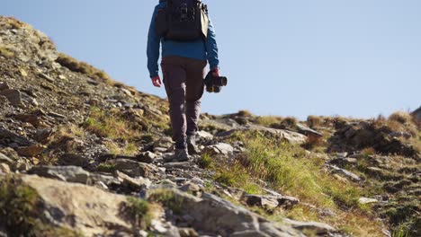 Solo-Male-Hiking-Content-Creator-Holding-Camera-Walking-Up-Rocky-Mountain-Hillside