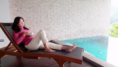 asian woman relaxing on a pool bed and drinking coffee by her private pool during her morning vacation