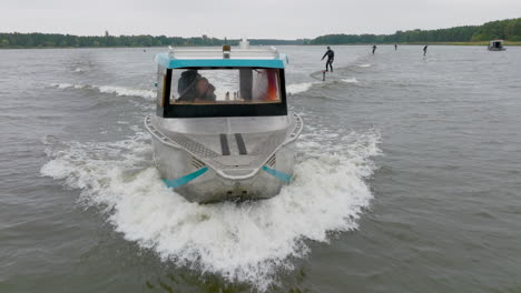 hydro foilers surfing wave behind watercraft driven by blond girl filmed with drone from front close up with trees on riverbank