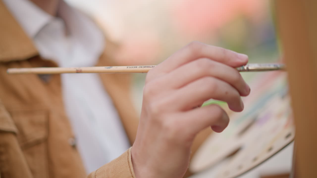 Nærbillede af maler, der påfører livlige grønne strøg på et lærred, med en palet i hånden og penselspidsen fuld af pigment. Kunstneren med jakke arbejder udendørs i en park og laver en energisk farveundersøgelse med livlige bevægelser.