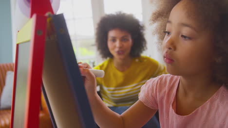 Mother-In-Wheelchair-Watching-Daughter-Having-Fun-Drawing-Picture-On-Whiteboard-At-Home