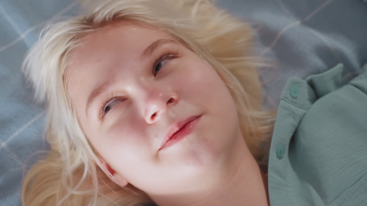 Close up of blonde kid with light hair smiling joyfully while singing and shaking body on soft blue bed, face lit with happiness and eyes nearly closed in delight during playful, carefree moment