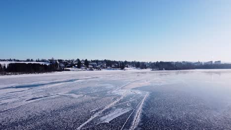 Ein-Ruhiger-Blick-Auf-Einen-Zugefrorenen-See-In-Norwegen,-Mit-Schneebedeckten-Häusern-Und-Bäumen-Im-Hintergrund-Unter-Einem-Klaren-Blauen-Himmel