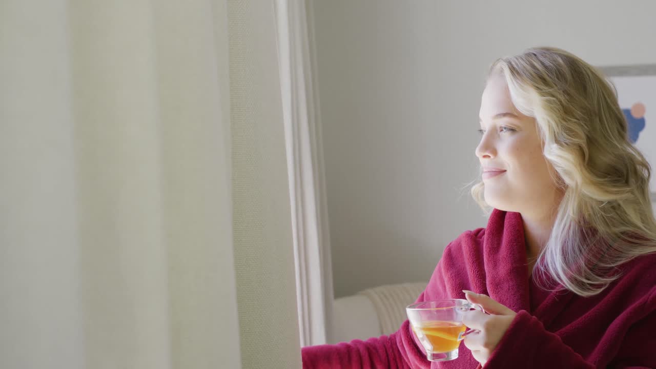 Happy caucasian plus size woman uncovering windows with curtain and drinking tea in slow motion