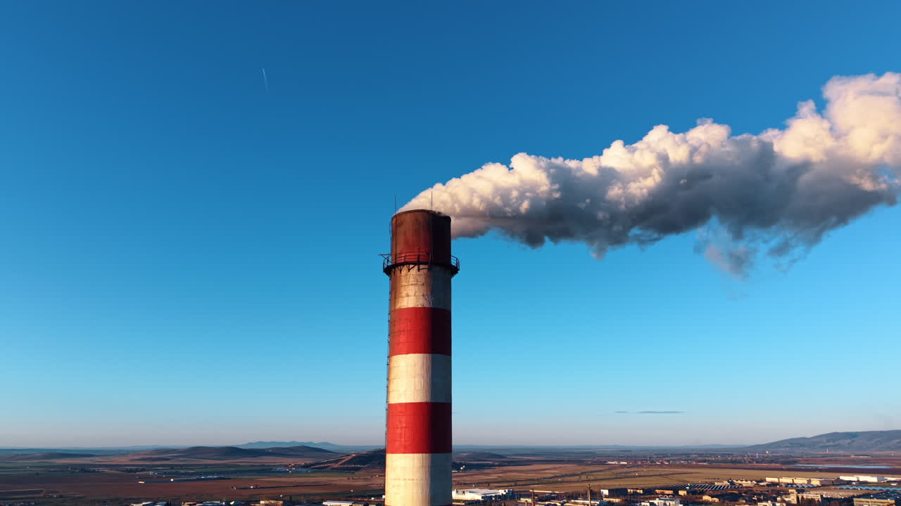 Ciminiera industriale con fumo su sfondo di cielo azzurro limpido. Ciminiera in cemento a strisce rosse e bianche che emette fumo di scarico bianco sopra la città e le montagne.