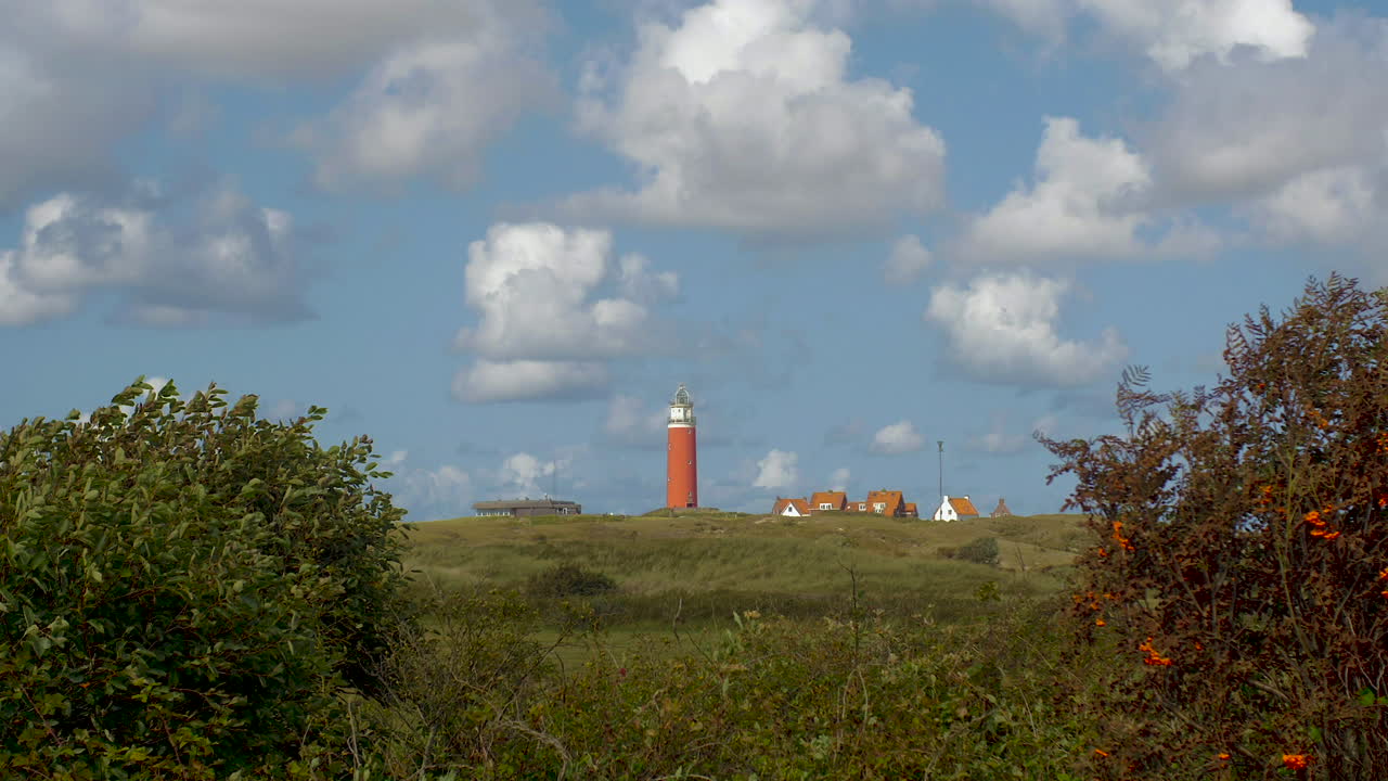 Time lapse through bushes in the foreground. In the distance we see a lighthouse in the dunes together with some builings. The sky is blue with beautiful clouds passing by.