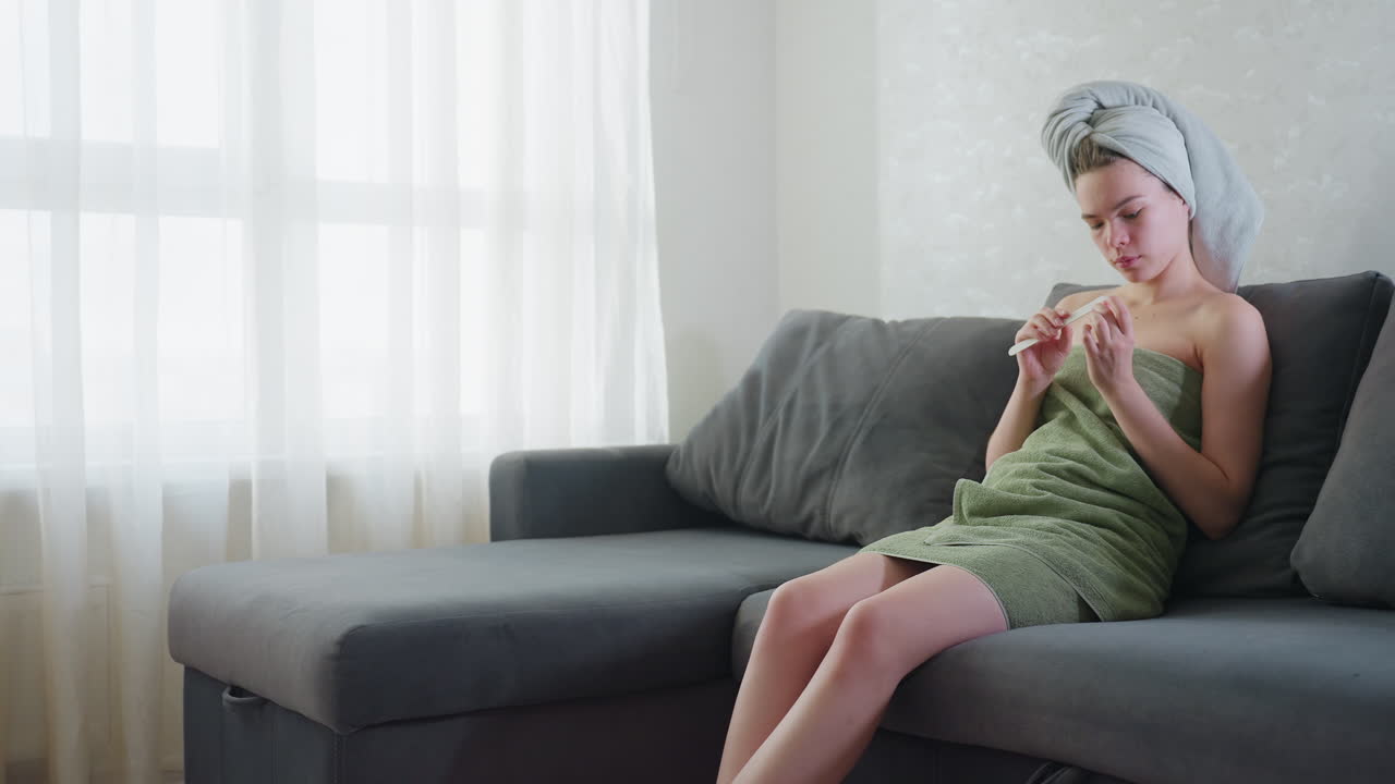 Young girl sits on grey couch in bright room wrapped in green towel with towel on head, focused on filing nails in peaceful atmosphere, soft daylight coming through sheer curtains