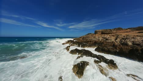 FPV-Racing-Drone-over-crystal-clear-blue-splashing-water-flying-towards-rocky-coast-line-shore-line