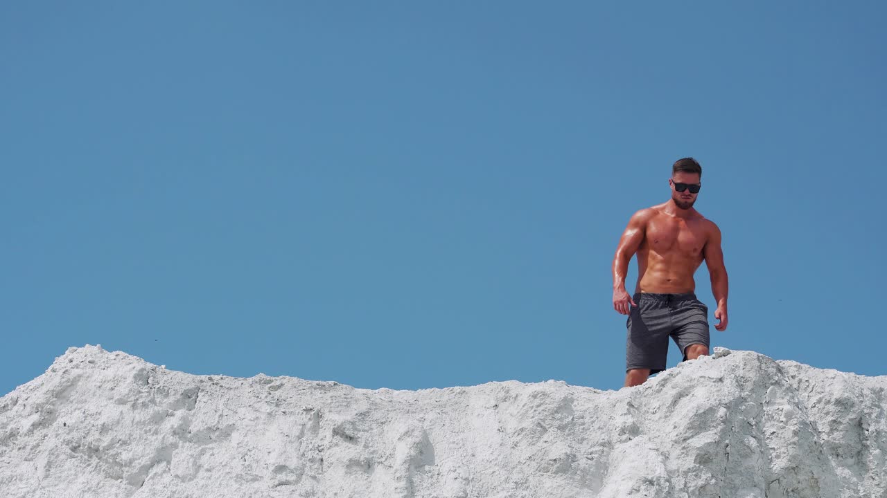 A shirtless muscular male bodybuilder runs along a white mountain against a blue sky