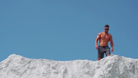 a shirtless muscular male bodybuilder runs along a white mountain against a blue sky